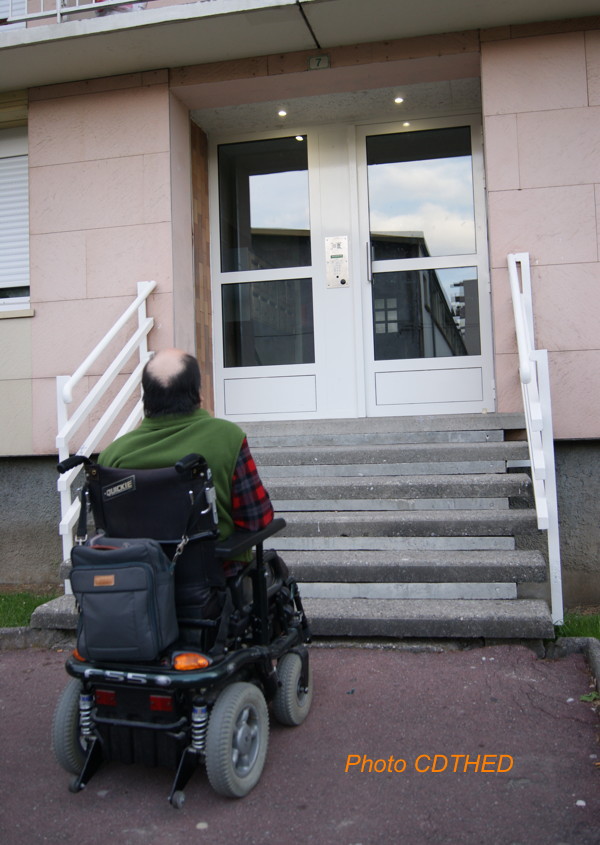 Sur la photo couleur, on voit de dos une personne handicapée en fauteuil roulant électrique. Devant elle, un escalier de six marches qui mène à une entrée d'immeuble... Inaccessible donc !